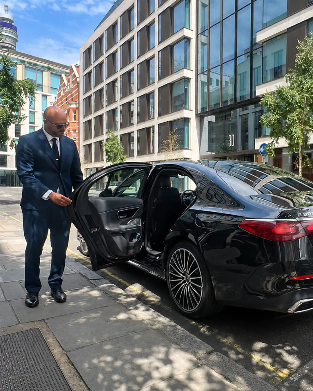 Chauffeur in a navy suit holding open the rear door of a black Mercedes outside a glass office building