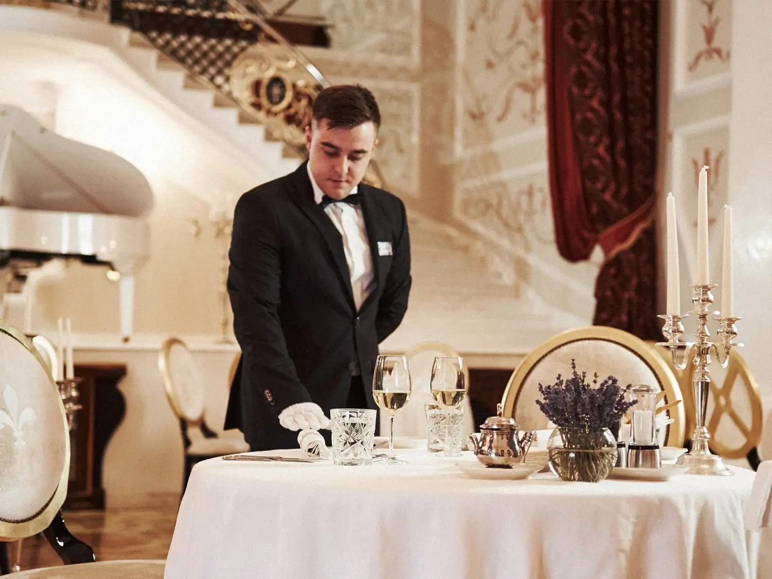 Butler in a black suit and white gloves arranging glassware at a formally set table in an ornate dining room