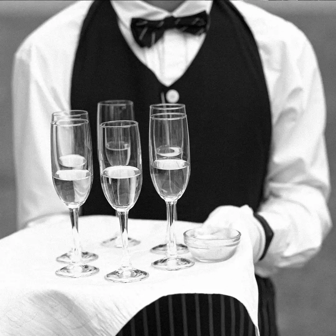 Butler in a waistcoat and bow tie holding a tray of champagne flutes, in black and white