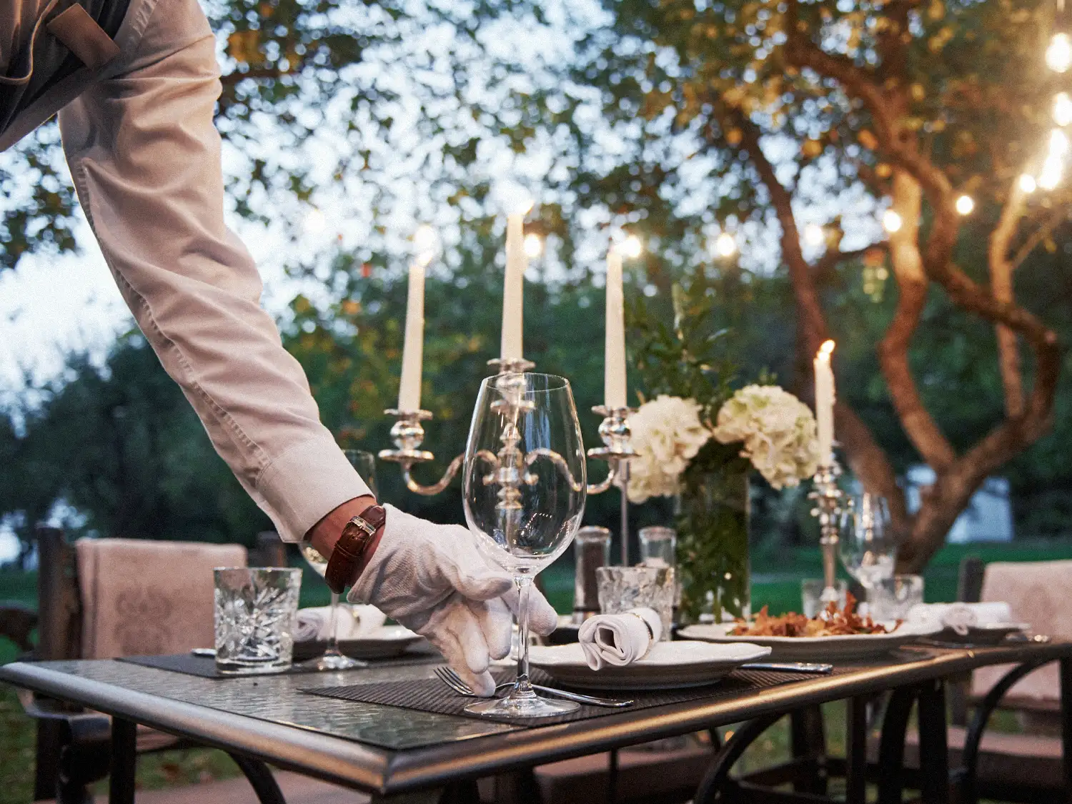 Butler in white gloves setting a candlelit outdoor dining table with crystal glasses and white flowers