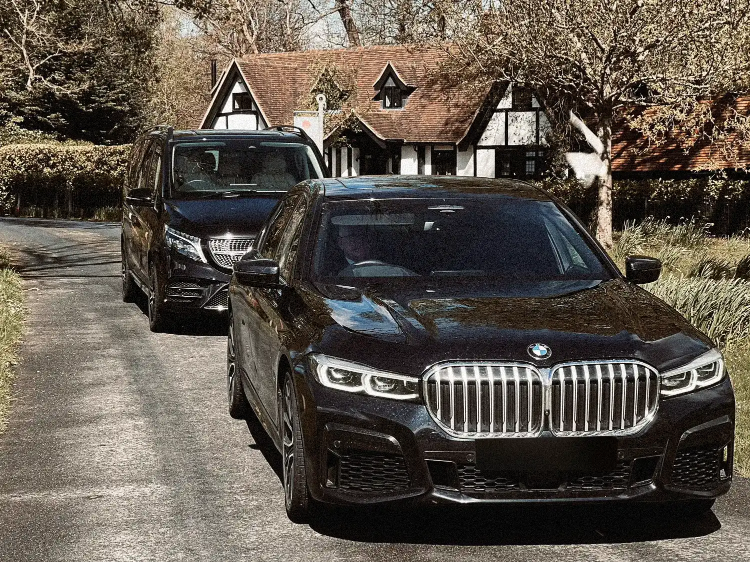 Black BMW 7 Series and executive people carrier parked outside a Tudor-style house for private chauffeur hire