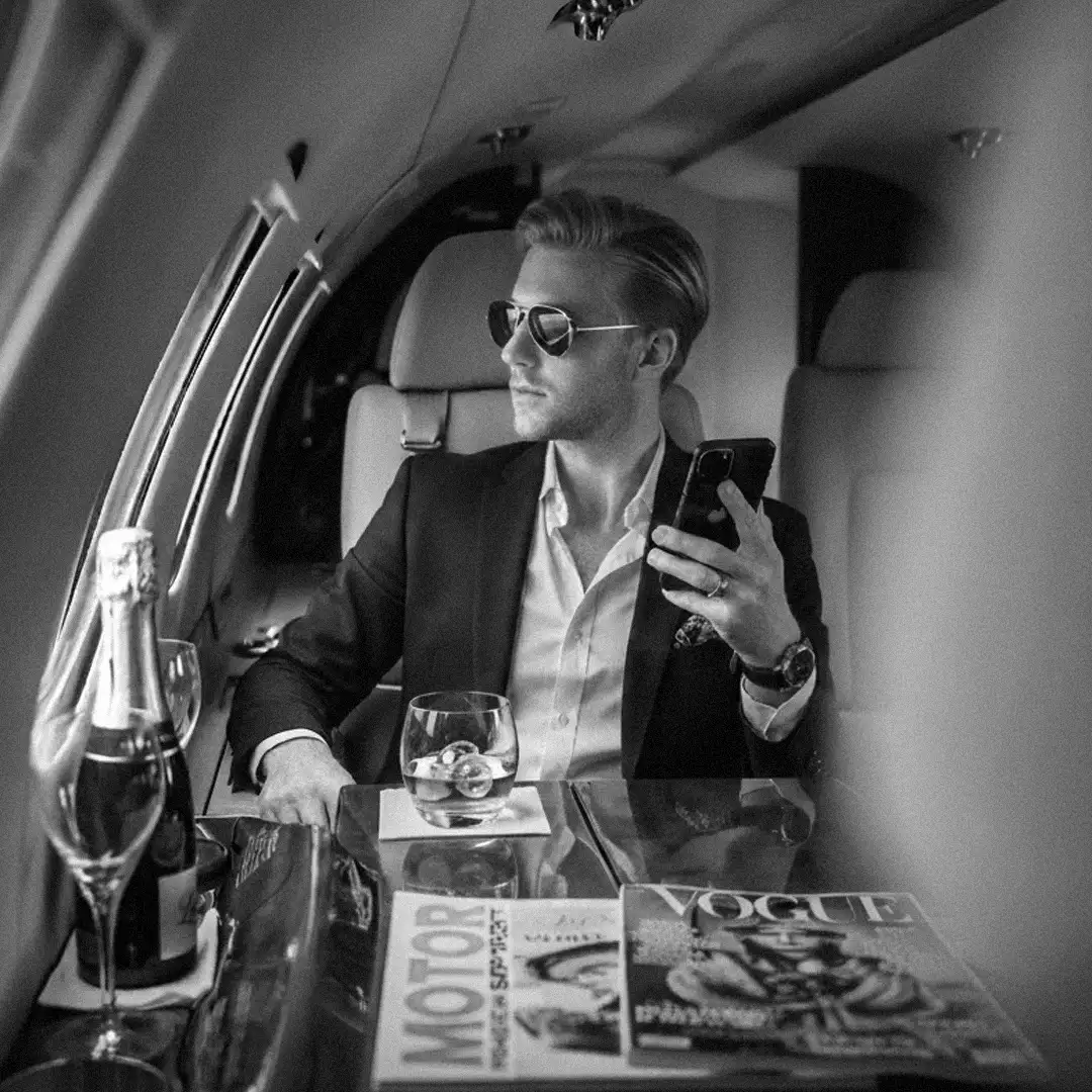 Man in sunglasses and a dark jacket checking his phone in a private jet cabin, with a champagne bottle and glass on the table